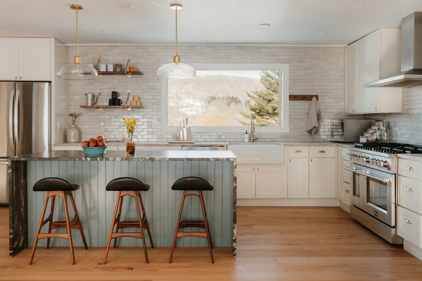 A prepared kitchen with a green island, brass fittings and a hillside view through the window